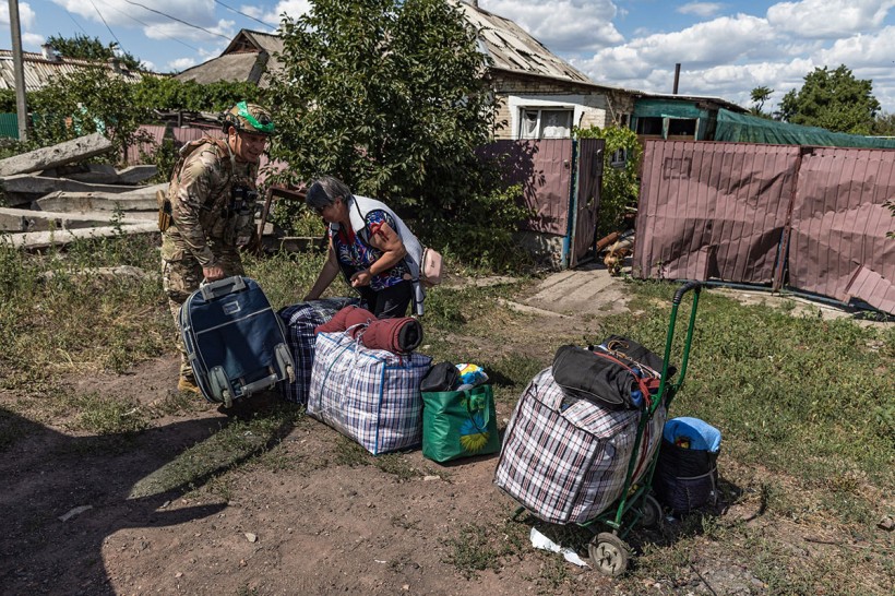 Жінка похилого віку стоїть на вулиці, біля неї великі сумки з речами, поруч поліцейський? який допомагає.