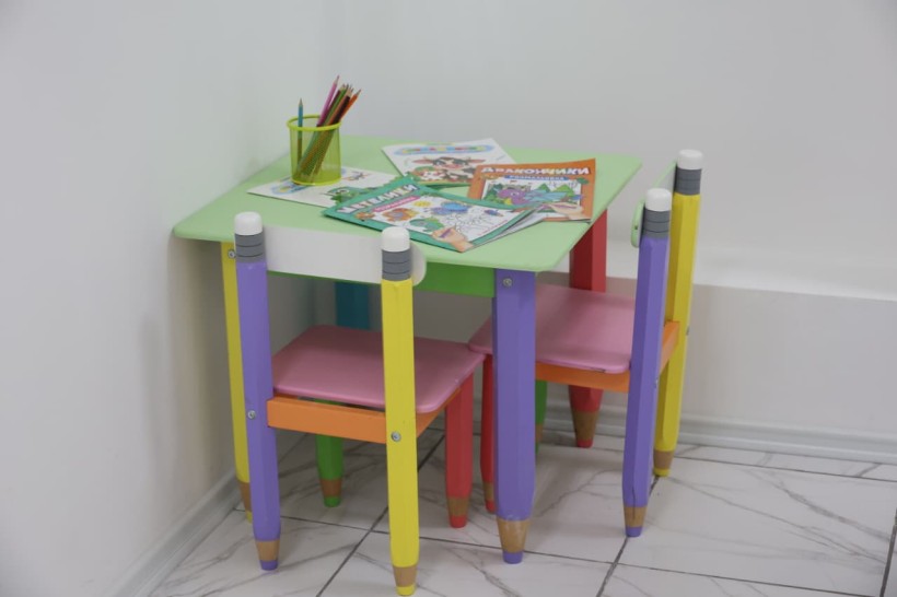 Дитячий куточок.
Різнокольоровий стіл та стільці. 
На столі лежать розмальовки та олівці.