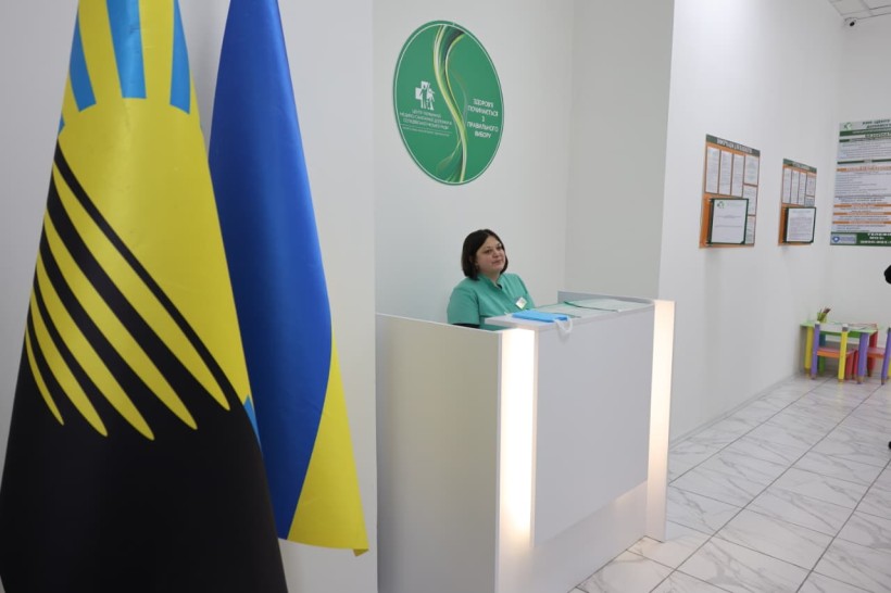 Хол медичного закладу.
Жінка адміністратор стоїть біля стійки приймання громадян.