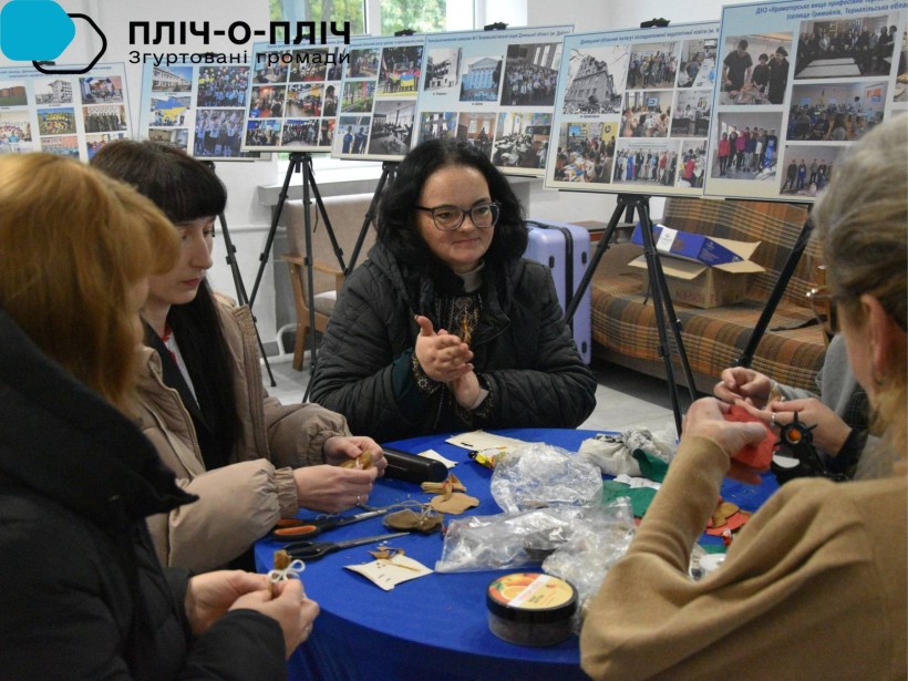 Жінки сидять за круглим столом, спілкуються та роблять сувеніри.