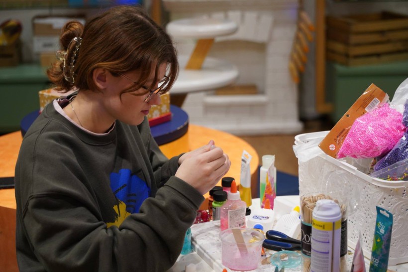 Дівчина в окулярах працює над поробом за столом, заставленим матеріалами для творчості.