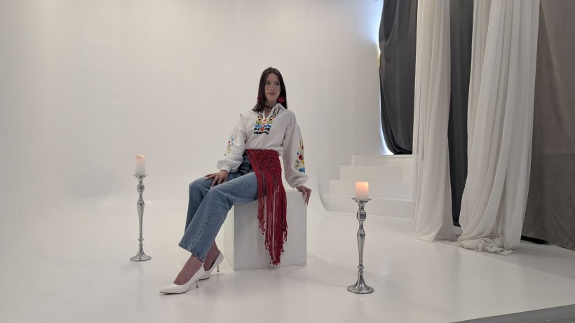Дівчина у вишиванці, джинсах та білих туфлях сидить на кубі у фотостудії між двома свічниками
