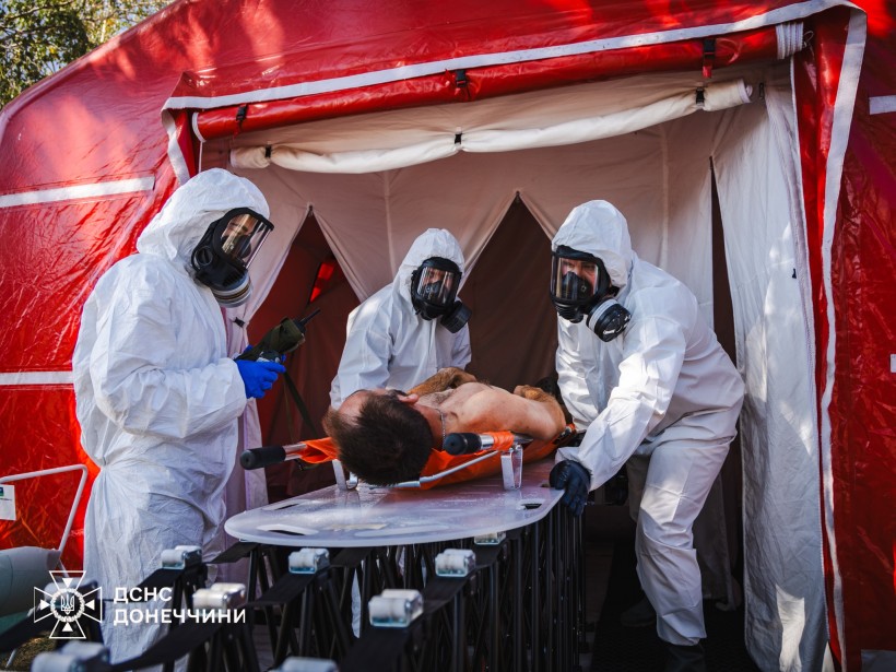 Рятувальники у спеціальних білих костюмах. Відпрацьовують техніку та допомогу постраждалим унаслідок хімічної атаки. На медичній кушетці лежить умовно потерпілий.