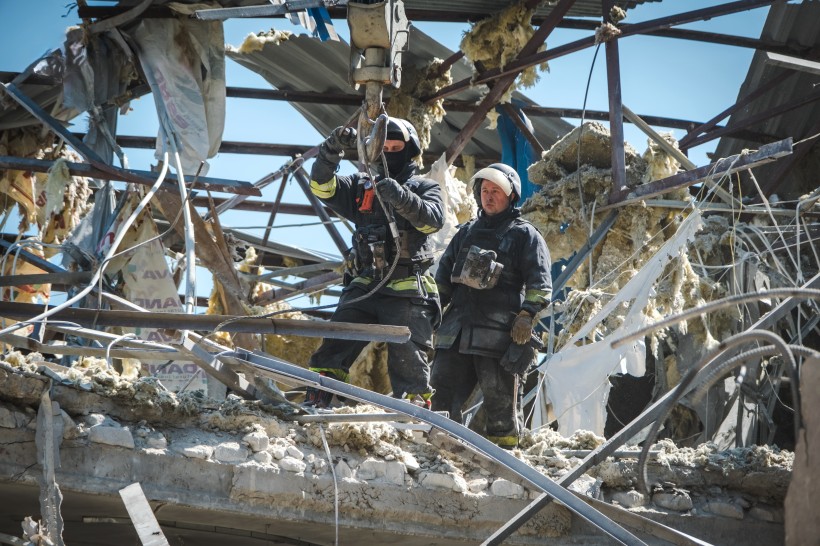 Двоє рятувальників у захисному спорядженні пробираються крізь уламки зруйнованої будівлі, оточені скрученим металом та ізоляційним матеріалом, під чистим небом.