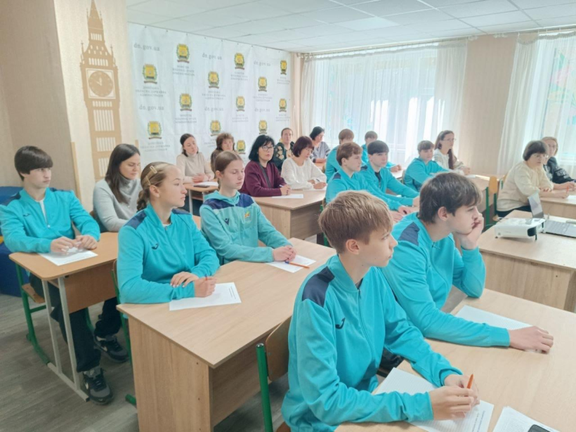 Молодь у формі та дорослі спортсмени сидять за партами в класі й пишуть диктант.