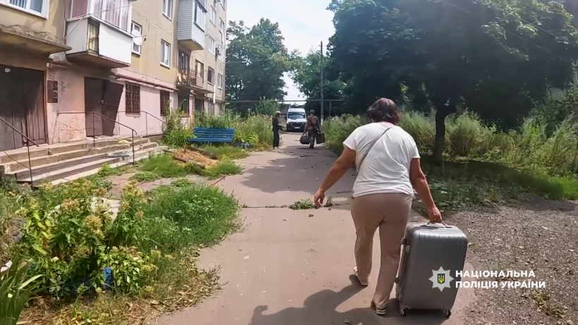 Жінка з валізою на колесах йде повз будинок, попереду - поліцейський з сумками, крокують на евакуацію