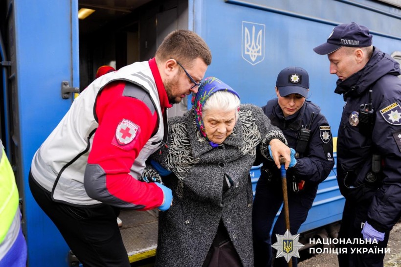 Представники поліції та Червоного Хреста допомагають літній жінці з палицею вийти з евакуаційного потяга