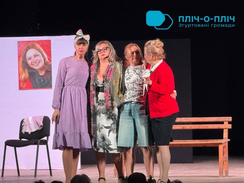 Чотири акторки стоять разом на театральній сцені в обіймах, виконуючи емоційну сцену; позаду видно лавку та фото на білому тлі