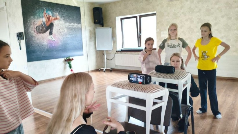 Заняття з хореографії: діти в залі показують танцювальні рухи для учасників, які повторюють їх онлайн через екран смартфона.