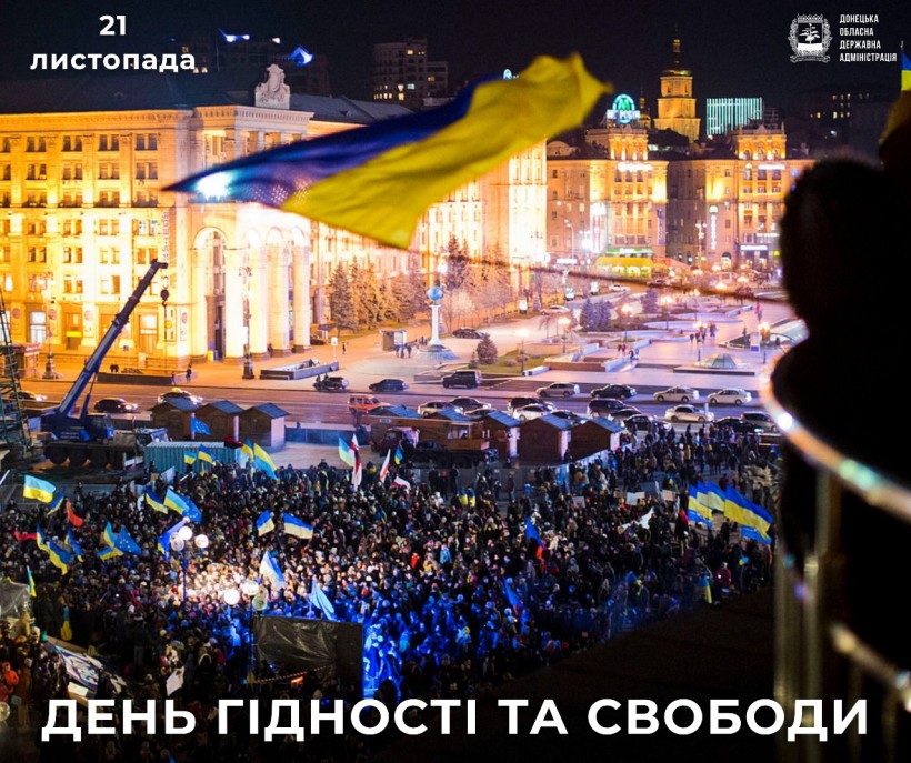 Натовп зібрався вночі на Майдані, тримаючи українські прапори. Будівлі яскраво освітлені. Текст українською мовою: «21 листопада» та «ДЕНЬ ГІДНОСТІ ТА СВОБОДИ».