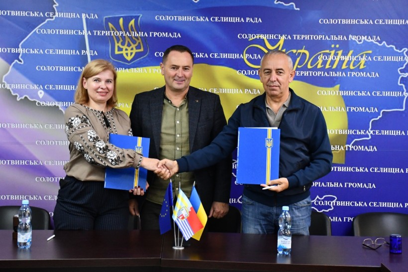 Троє людей стоять і тиснуть один одному руки на тлі з українськими мотивами. Вони тримають сині папки, що символізують угоду або партнерство. На столі лежать маленькі прапорці.