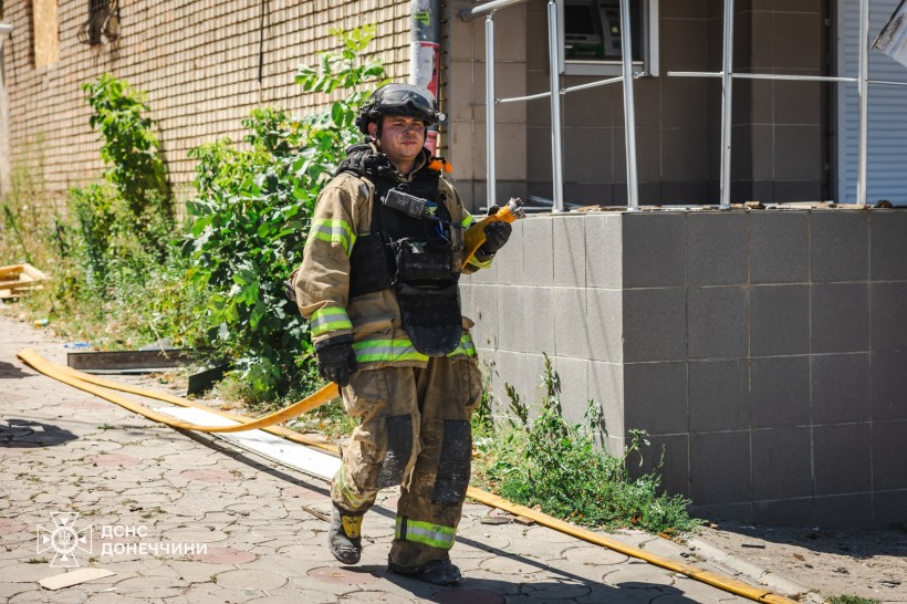 Рятувальник несе у руках гідравлічний шланг. З боку нього понівечена будівля. Одягнений у шолом та броніжелет