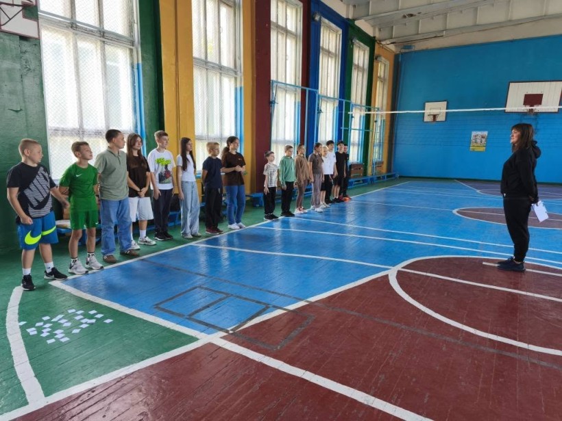 Спортивний зал. Діти стоять шеренгою у спортивному одязі. По центру стоїть жінка у чорному костюмі та щось розповідає.