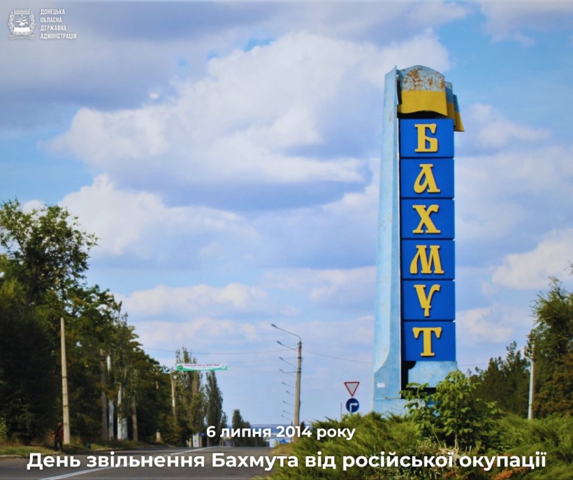 Високий синій знак з жовтим написом «БАХМУТ» на дорозі, обсадженій деревами, під чистим блакитним небом. Напис: «День визволення — Бахмут, 6 липня 2014 року».