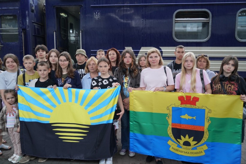 Діти та дорослі стоять на пероні вокзалу. Позаду них вагон потяга синього кольору. На передньому плані діти тримають прапори громади та Донеччини.