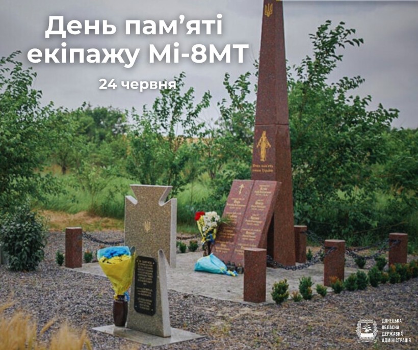 Урочисте меморіальне місце з високим червоно-коричневим обеліском, оточеним зеленню. Біля його основи покладені квіти.