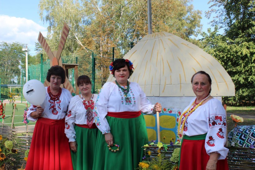 Жінки у національному вбранні, зелені та червоні спідниці, зверху одягнені вишиванки та намисто. Поруч макет української хати