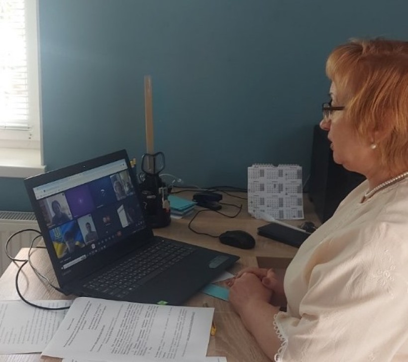 Заступниця начальника Іллінівської сільської військової адміністрації Наталія Лебедева, яка сидить за столом і дивиться в ноутбук, на екрані якого видно кілька учасників відеодзвінка. Перед жінкою на столі лежать документи.