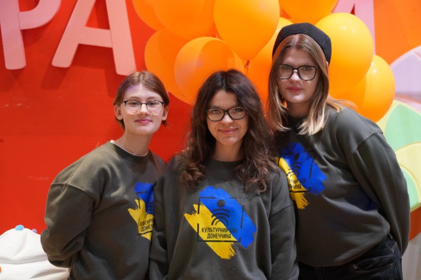 Три дівчини в однакових оливкових світшотах з написом «Культурний хаб Донеччина» на фоні помаранчевих кульок.
