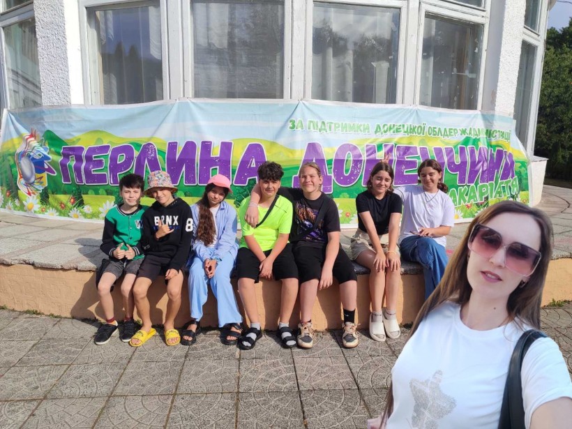 Діти сидять біля фасаду будівлі, на якому висить банер з надписом "Перлина Донеччини", жінка робить селфі.