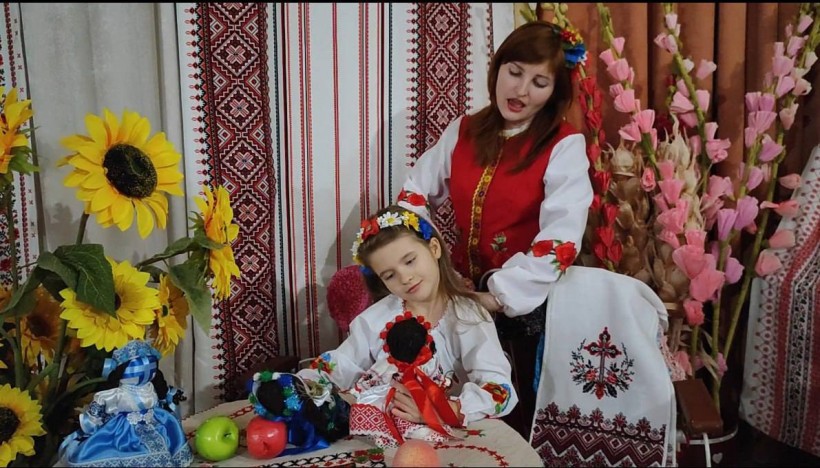 Мати з дочкою за столом співають, тримаючи ляльки мотанки у руках