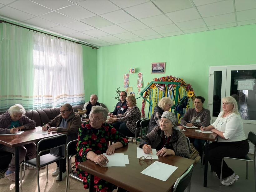 Літні люди разом з іншими учасниками сидять за столами та пишуть диктант у кімнаті.