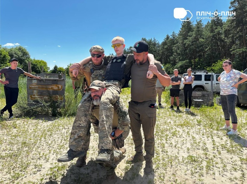 Інструктори та учасники практичного курсу з тактичної медицини тримають учасника курсу за руки, третій інструктор підтримує його тазову частину своєю спиною, сидячі на колінах
