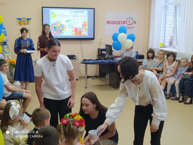 Урочистий захід до Дня знань у приміщенні освітньої локації Курахівської ТГ м. Дніпро: випускники вітають першокласників. Навколо — кульки, глядачі та святкова атмосфера.