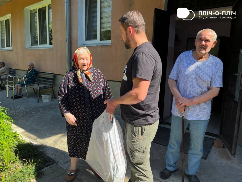 Заступник начальника Часовоярської громади стоїть біля жінки та чоловіка похилого віку. В руках тримає пакет з гуманітарною допомогою. Жінка щось розповідає.