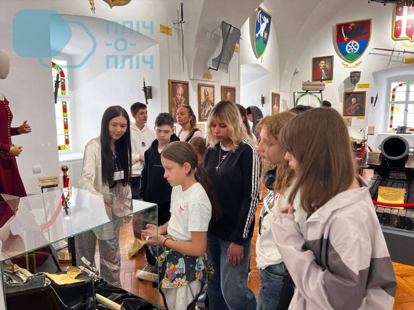 Хлопці та дівчата уважно роздивляються предмети старовини. На стінах портрети гетьманів та герби.