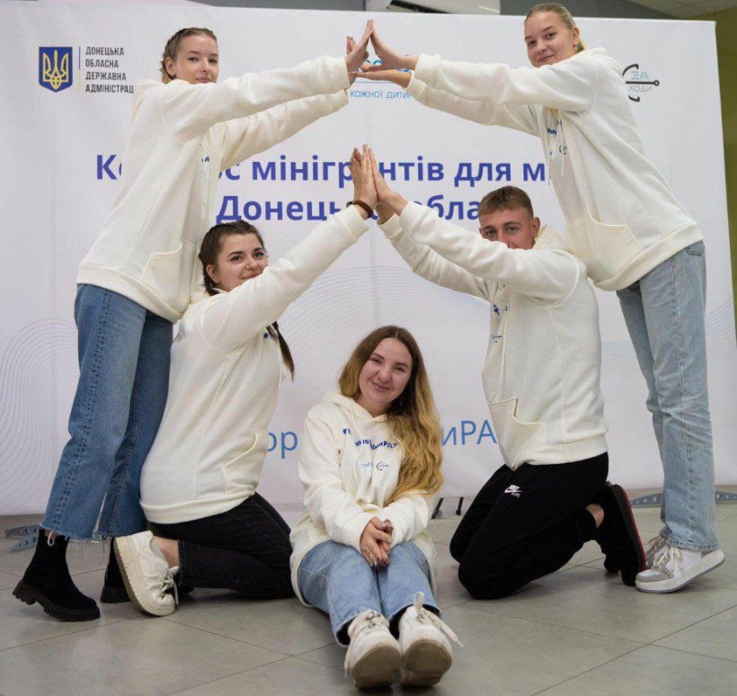 П’ятеро молодих людей у білих худі утворюють фігуру у формі будинку, тримаючи долоні разом над головою дівчини, яка сидить посередині, під час молодіжного заходу для молоді Донеччині.