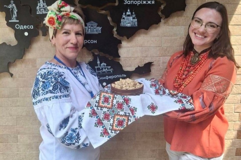 Жінки у вишиванках тримають глиняну миску зі стравою на вишитому рушнику на тлі декоративної карти України