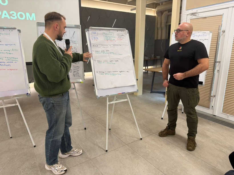 Обговорення робочих процесів.  Молодий чоловік з мікрофоном щось розповідає та показує рукою на мольберт, поруч стоїть начальник міської військової адміністрації Сергій Чаус.