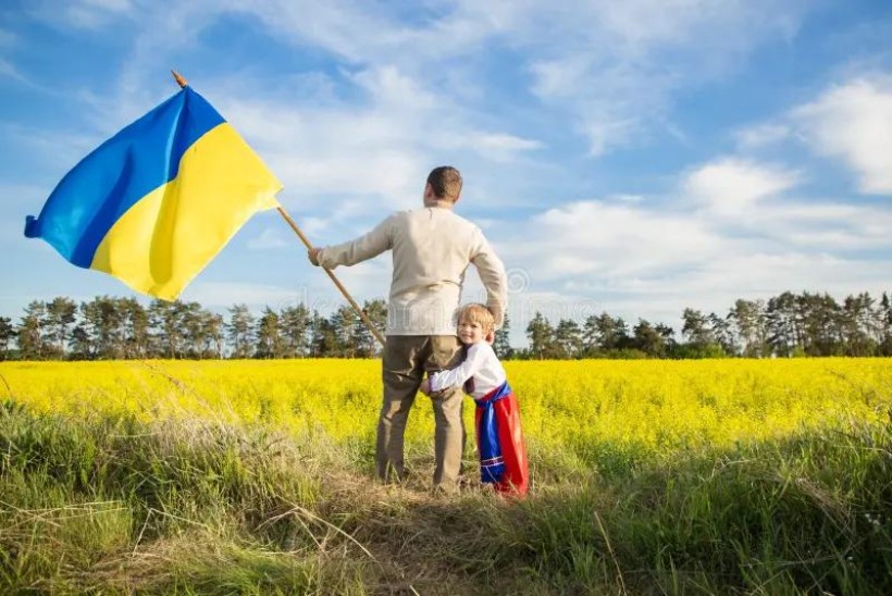 Чоловік тримає прапор України, його обіймає маленький хлопчик, обидва стоять у полі