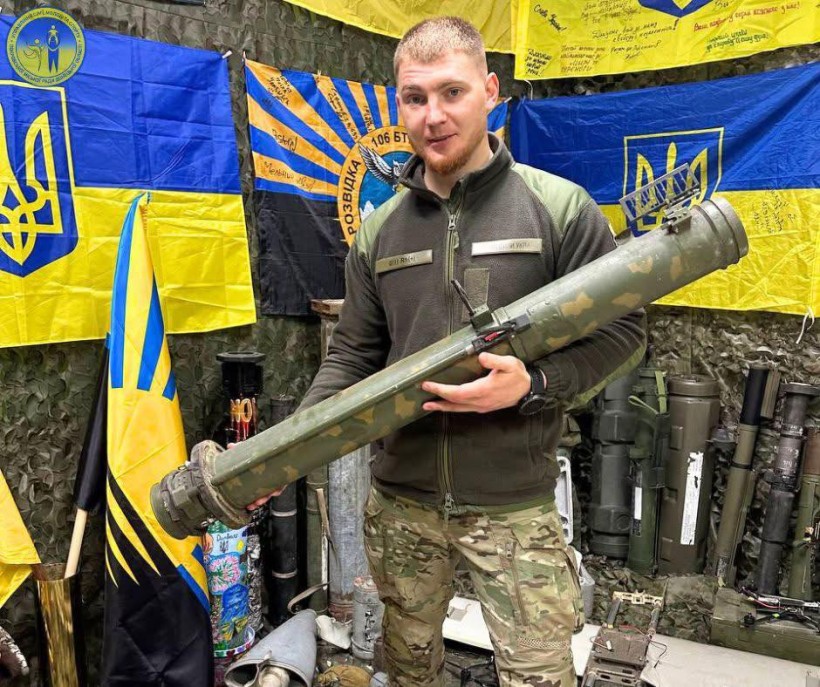 Військовий тримає на руках використаний тубус від снаряда. Поруч прапори військових частин.