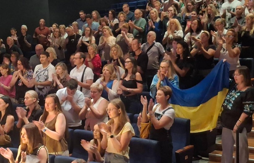 Глядачі у залі театру аплодують стоячи, жінка в першому ряду тримає на плечах прапор України.