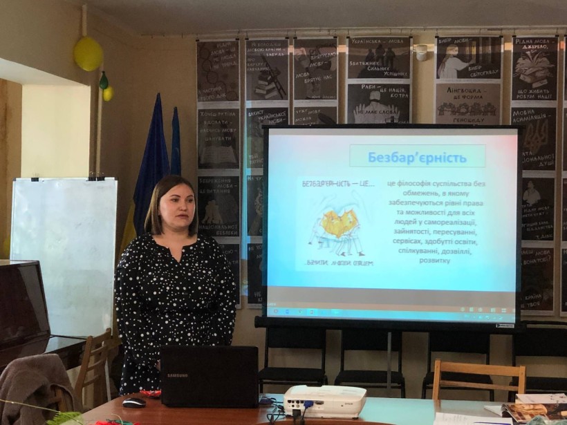 Лекторка стоїть поруч із ноутбуком на фоні екрана, на якому відображено слайд із заголовком «Безбар’єрність»