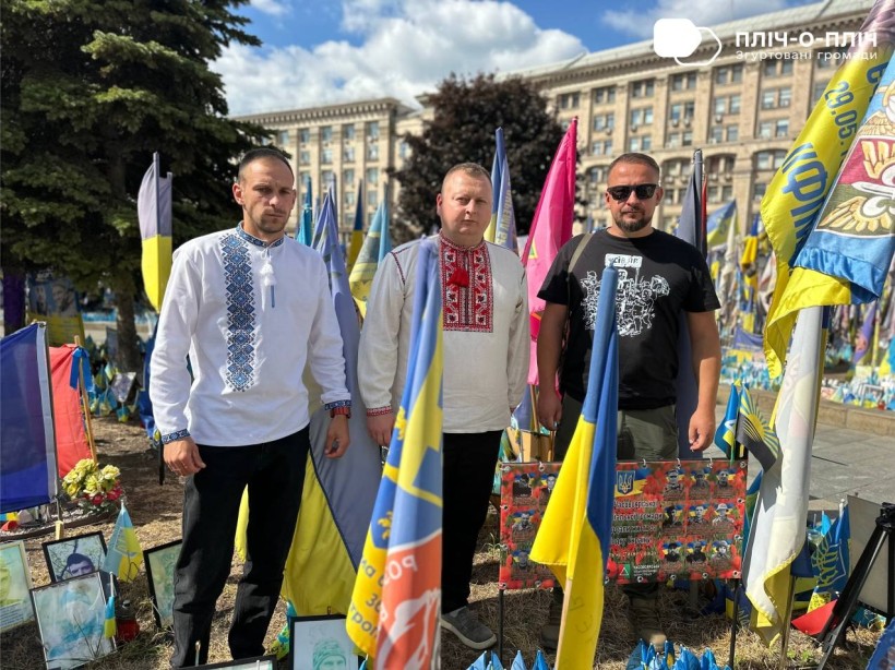 Троє чоловіків у вишиванках стоять на алеї Слави у місті Київ. Поруч прапор військових бригад та частин.