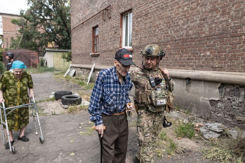 Поліцейський допомагає літньому чоловіку евакуюватись з міста Костянтинівка. Веде чоловіка під руку. Позаду літня жінка з ходунками