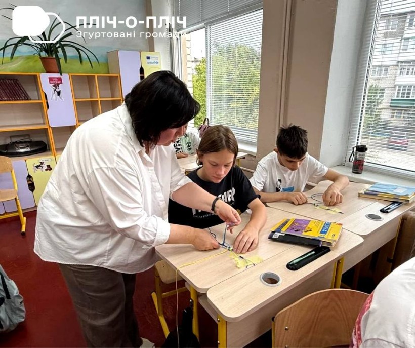 Двоє учнів сидять за партами та роблять захисні амулети для захисників, вчитель поруч допомагає.