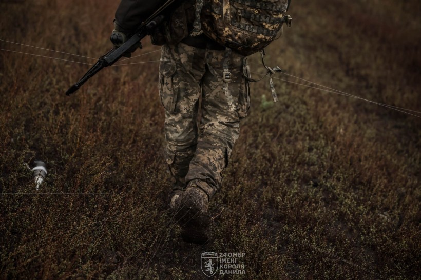 Поле. Розтягуйте оптоволокно, поруч на землі лежить ворожий дрон. Військовий зі зброєю стоїть спиною.