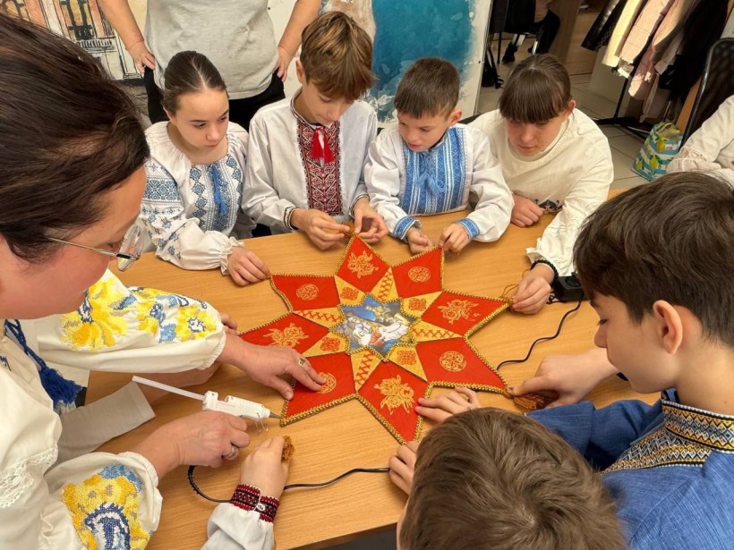 Діти у вишиванках разом із дорослим виготовляють велику восьмикутну різдвяну зірку з червоної тканини, оздоблену золотистими візерунками.