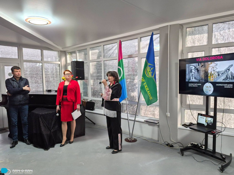 Жінка у мікрофон розповідає про виставку, позаду прапори громад.