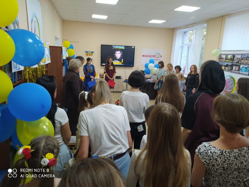 Учасники заходу стоять під час хвилини мовчання, вшановуючи пам’ять загиблих. На екрані зображення військового. Приміщення прикрашене синьо-жовтими кульками.