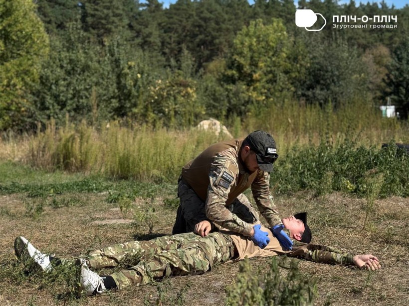 Двоє чоловіків у воєнній формі. Один з них лежить на землі другий показує вправи з тактичної медицини.