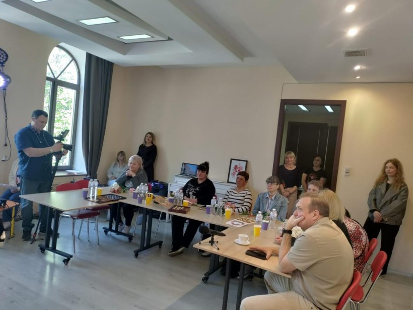 Матері Героїв та гості заходу сидять за столами в залі, чоловік з відеокамерою знімає захід