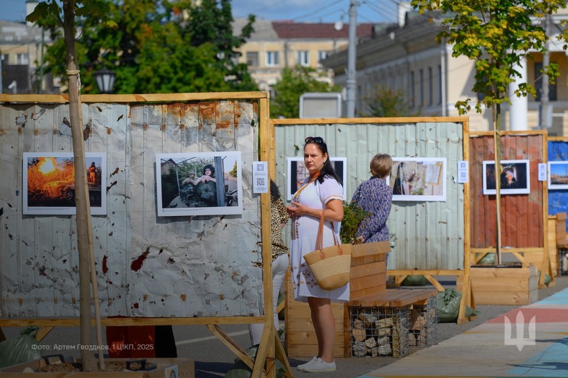 Молода жінка у світлій сукні уважно дивиться представлені фотографії війни. Позаду неї ще одна відвідувачка. Фотографії розвішані на металевих площах.