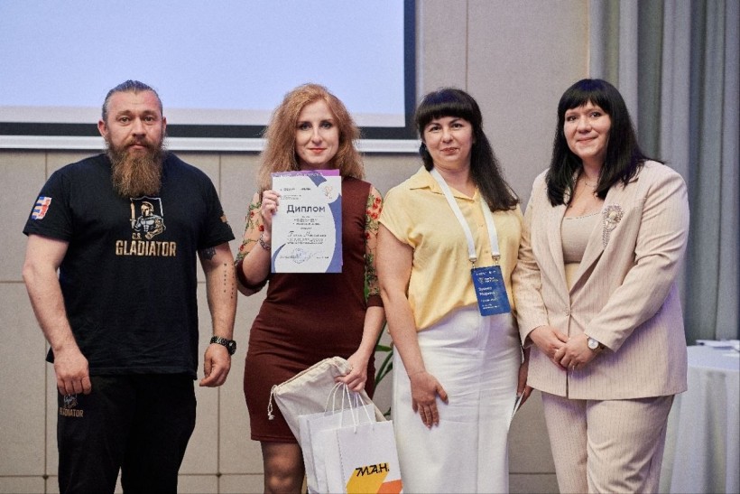 Церемонія нагородження: чотири людини на сцені, Ганна Никитська в центрі тримає диплом, поруч колеги та подарунковий пакет з логотипом МАН.