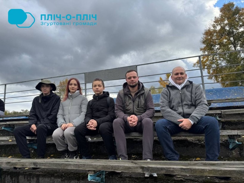 Група з п'яти осіб сидить на відкритих трибунах під хмарним небом. Вони одягнені в куртки, що свідчить про прохолодну погоду. Настрій здається спокійним і невимушеним.