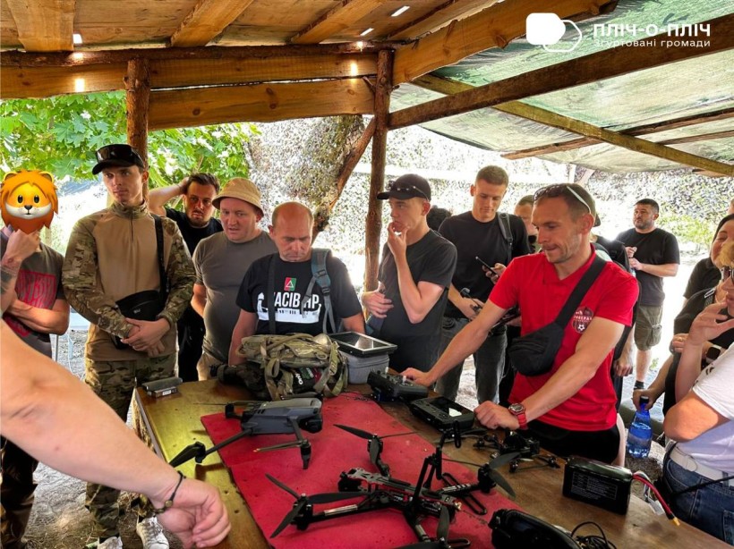 Чоловіки стоять біля столу. На столі лежать безпілотні апарати. Інструктор розповідає про їх дію та характеристики. Всі слухають.